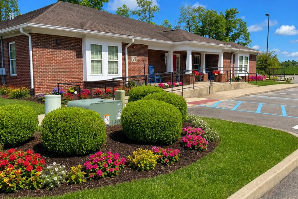 Welcoming red brick Shepherd's House long-term recovery housing home with a white-columned porch, accessibility ramp, and vibrant flower gardens